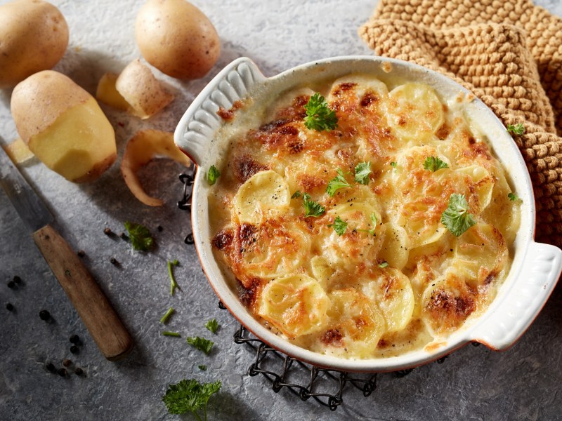 Eine runde Auflaufform mit Kartoffelgratin mit Pilzen, daneben teilweise geschälte Kartoffeln und ein Messer sowie Kräuter.