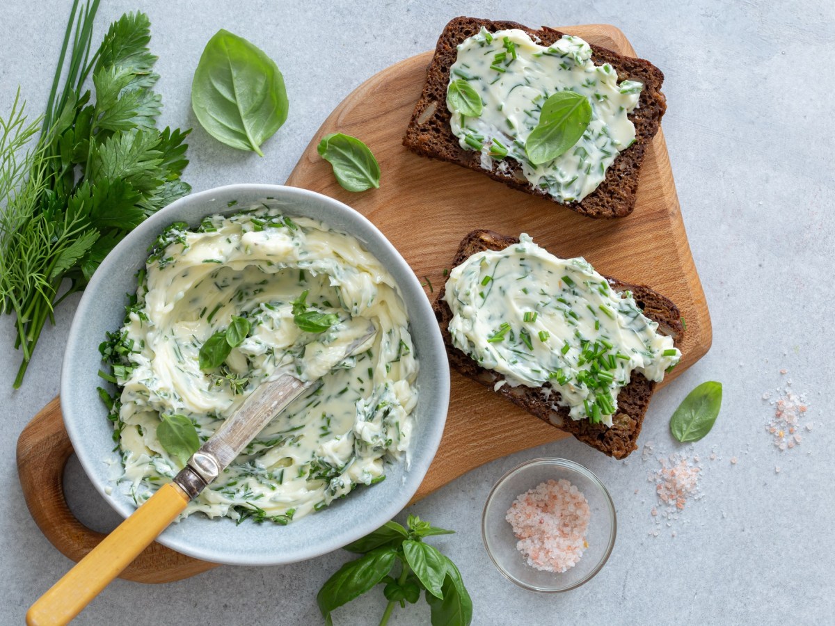 Eine Schüssel Knoblauch-Parmesan-Butter auf einem Brett, auf dem auch zwei Brotscheiben mit der Butter bestrichen liegen, daneben frische Kräuter und ein Schälchen Salz. Draufsicht.