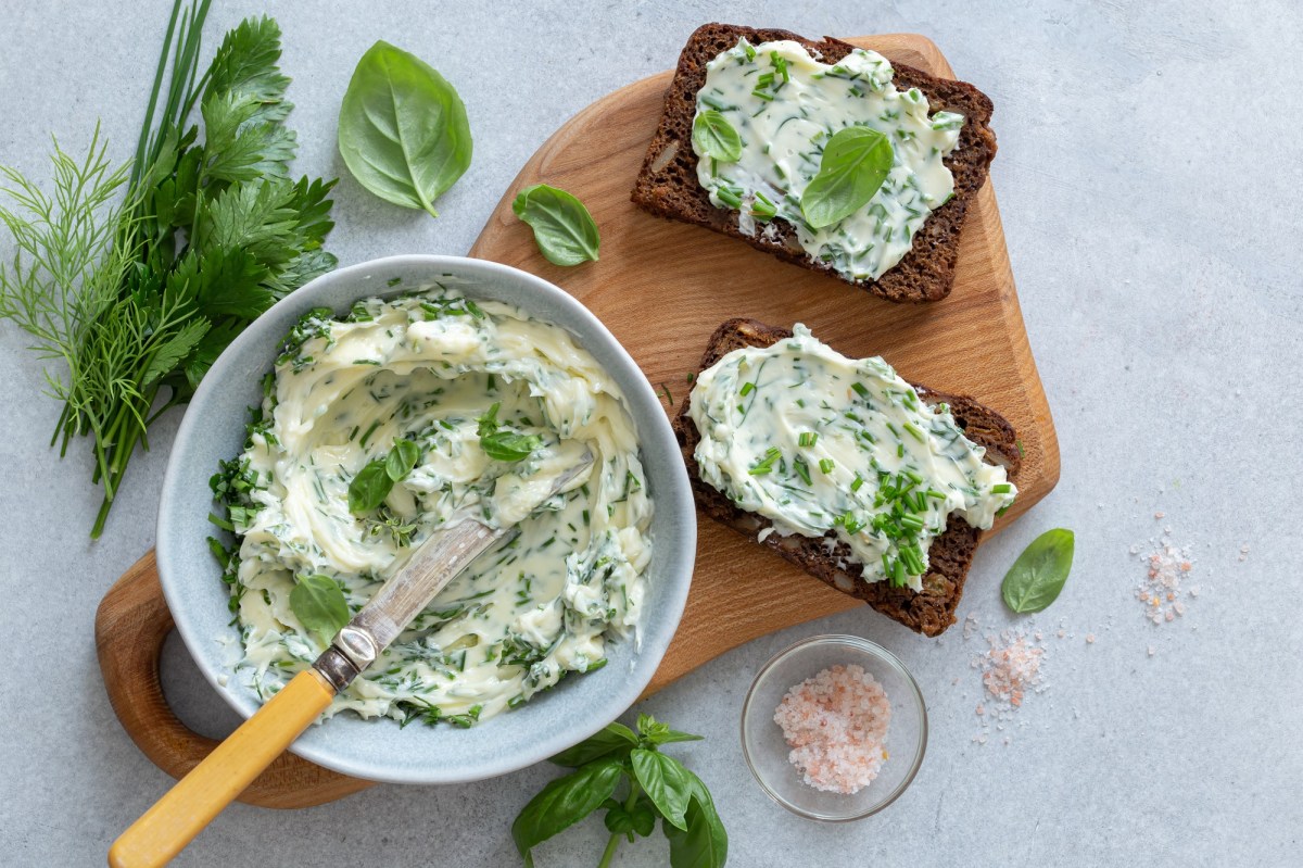 Eine Schüssel Knoblauch-Parmesan-Butter auf einem Brett, auf dem auch zwei Brotscheiben mit der Butter bestrichen liegen, daneben frische Kräuter und ein Schälchen Salz. Draufsicht.