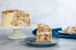 Ein Stück Kolibri-Kuchen auf einem Teller, im Hintergrund der angeschnittene Kuchen mit weißer Glasur verziert.