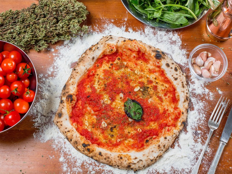 Pizza Marinara mit Tomatensoße, Knoblauch und einem Blatt Basilikum in der Draufsicht, daneben eine Schale Tomaten, getrockneter Oregano, Basilikum und Besteck,