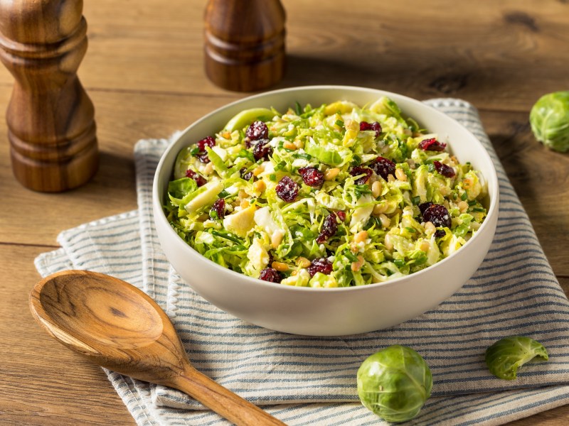 Eine weiße, flache Schale, in der sich Rosenkohlsalat mit Cranberrys und Pinienkernen befindet.