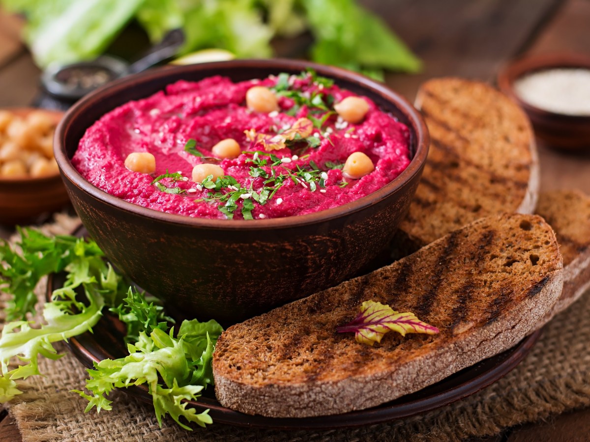 Rote Bete Hummus in einer dunklen Schüssel. dekoriert mit Kichererbsen und Kräutern. Serviert wird es mit Brot.