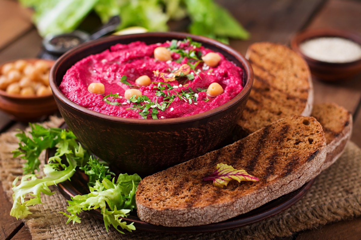 Rote Bete Hummus in einer dunklen Schüssel. dekoriert mit Kichererbsen und Kräutern. Serviert wird es mit Brot.