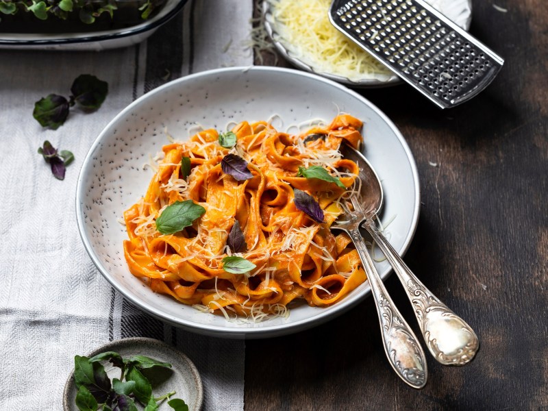 Ein Teller mit Paprika-Pasta mit Käse und Basilikum garniert, darauf Besteck, daneben eine Schale mit Käse und eine Reibe.