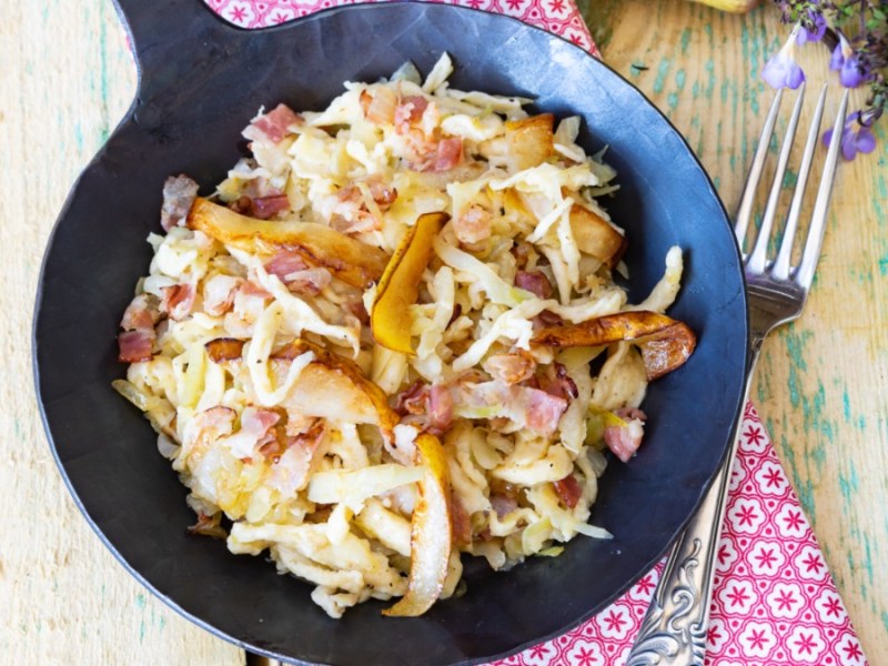 Eine Schupfnudel-Krautpfanne mit Birne und Speck von oben fotografiert. Daneben liegt eine Gabel.