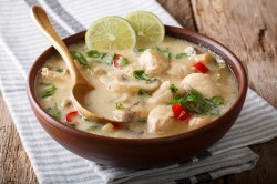 Tom Kha Gai Suppe in einer braunen Schale mit einem Holzlöffel. Sie ist mit zwei Limettenscheiben dekoriert.