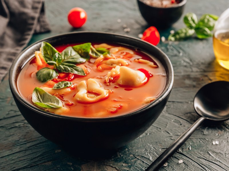 Eine schwarze Schüssel Tortellini-Suppe mit roter Tomatensuppe und Basilikumblättern, daneben ein Löffel, eine Flasche mit Öl und wenige weitere Zutaten.