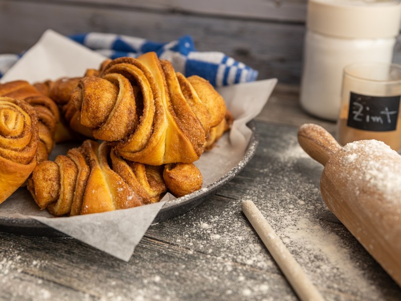 Ein paar vegane Franzbrötchen auf einem tiefen Teller, daneben ein Nudelholz, ein Glas mit Zimt und eins mit Zucker.