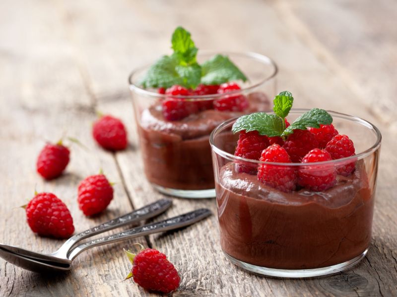 Vegane Mousse au Chocolat, serviert in zwei Gläsern. Himbeeren dienen als Deko in den Gläsern und daneben.