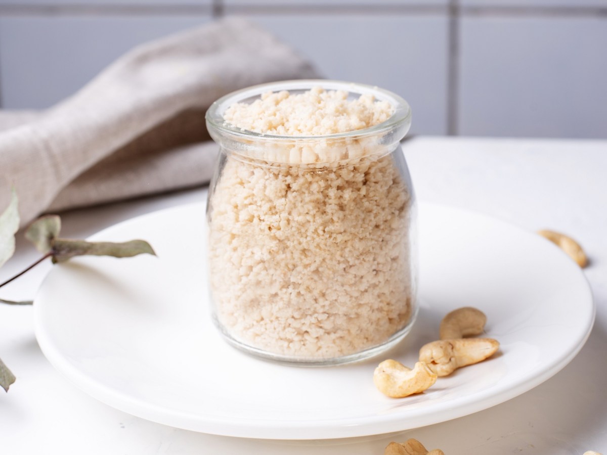 Ein Glas veganer Parmesan aus Cashews auf einem Teller, daneben ein paar Cashews.