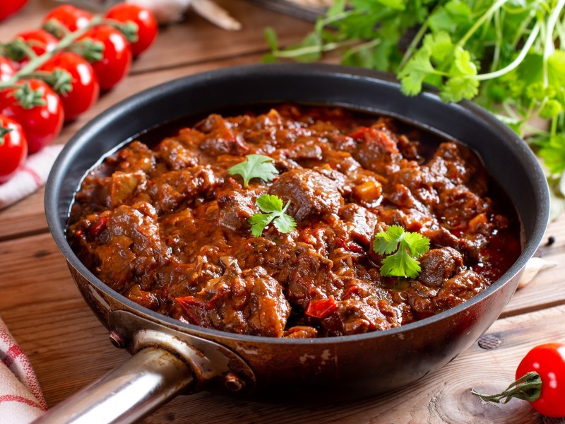 Eine Pfanne veganes Gulasch mit Möhren und Petersilie bestreut, drumherum Rispentomaten und frische Kräuter.