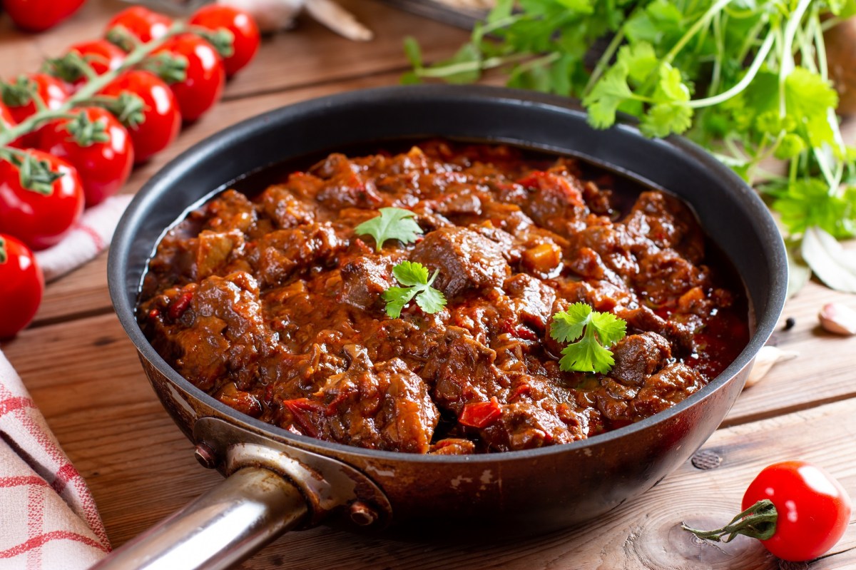 Eine Pfanne veganes Gulasch mit Möhren und Petersilie bestreut, drumherum Rispentomaten und frische Kräuter.