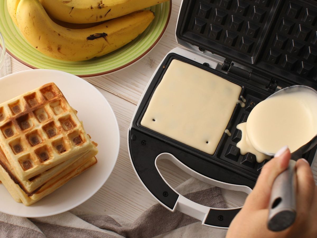 Ein Waffeleisen wird mit Teig gefüllt, eine zweite Waffel backt schon im Eisen. Daneben steht ein Teller mit gebackenen Waffeln. Im Hintergrund sind Bananen zu sehen.