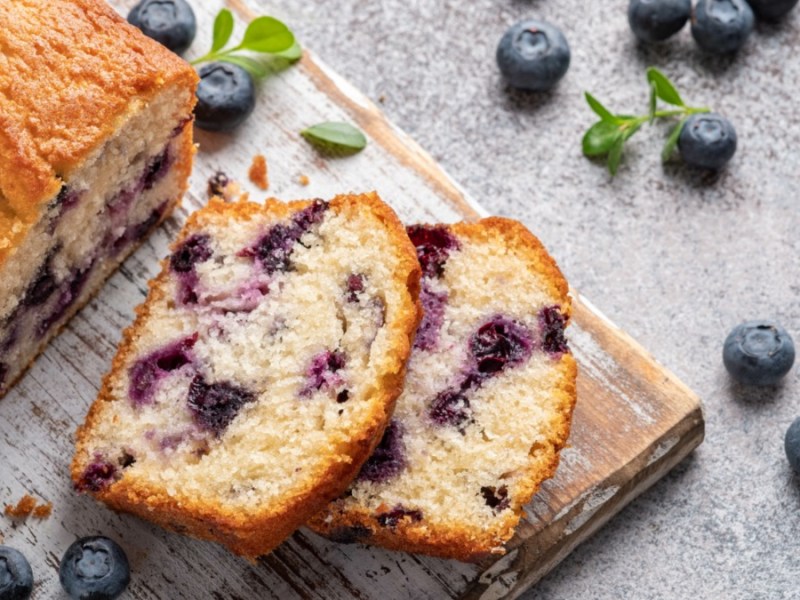 Ein Zitronen-Blaubeer-Brot auf einem Brett, von dem zwei Scheiben abgeschnitten wurden und daneben liegen. Daneben liegen frische Blaubeeren und Minzblätter.