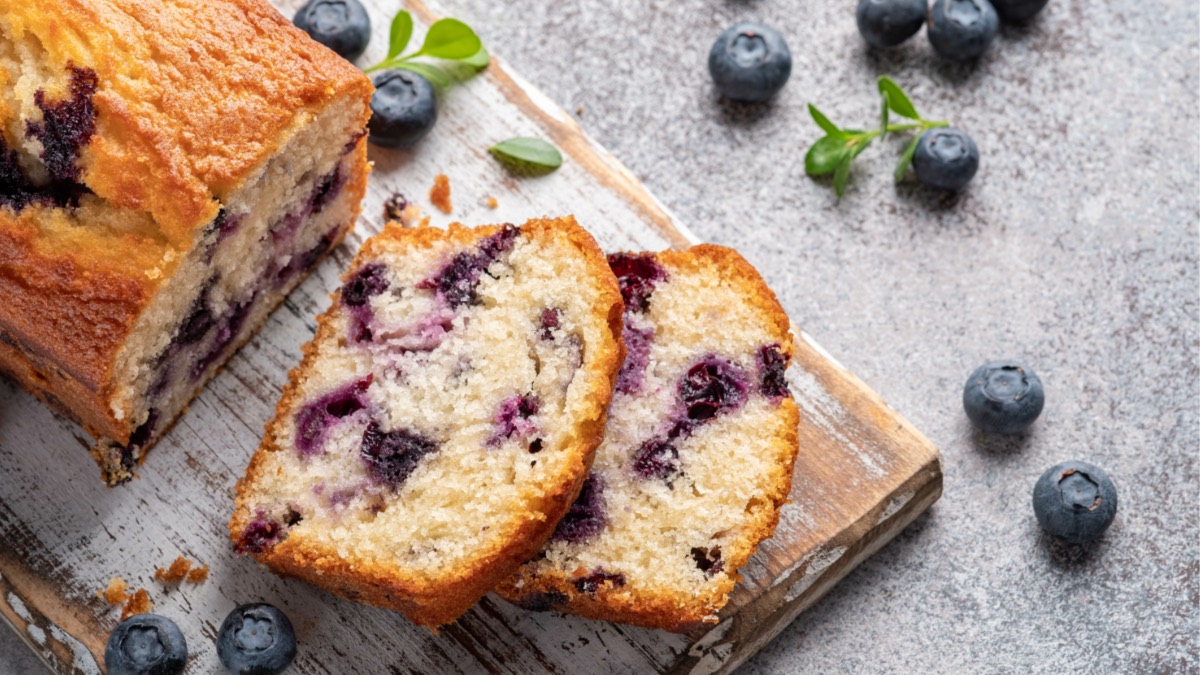 Ein Zitronen-Blaubeer-Brot auf einem Brett, von dem zwei Scheiben abgeschnitten wurden und daneben liegen. Daneben liegen frische Blaubeeren und Minzblätter.