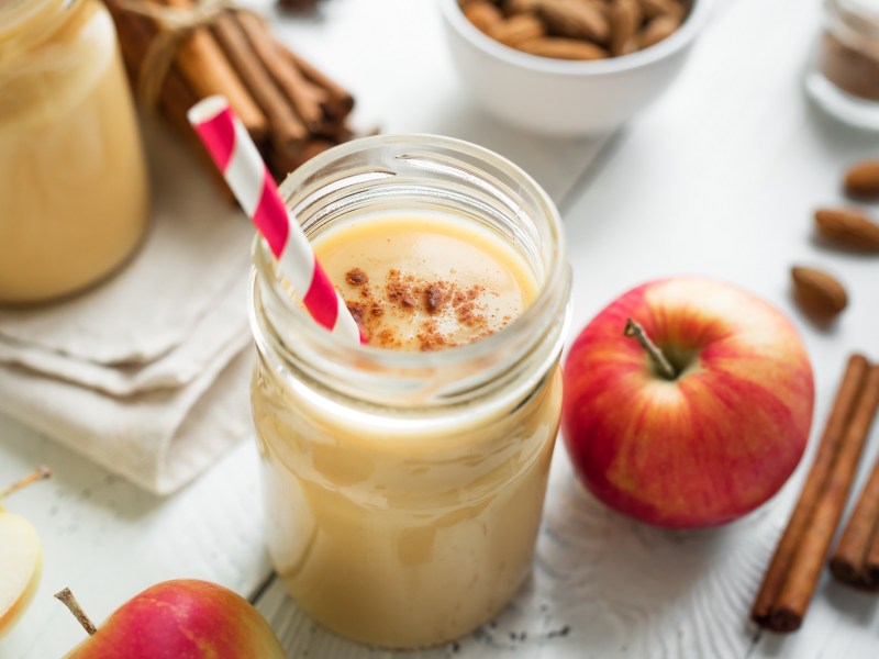 Ein Glas Apfel-Bananen-Smoothie mit Zimt bestreut, darin ein Strohhalm, drumherum Zimtstangen und Äpfel.