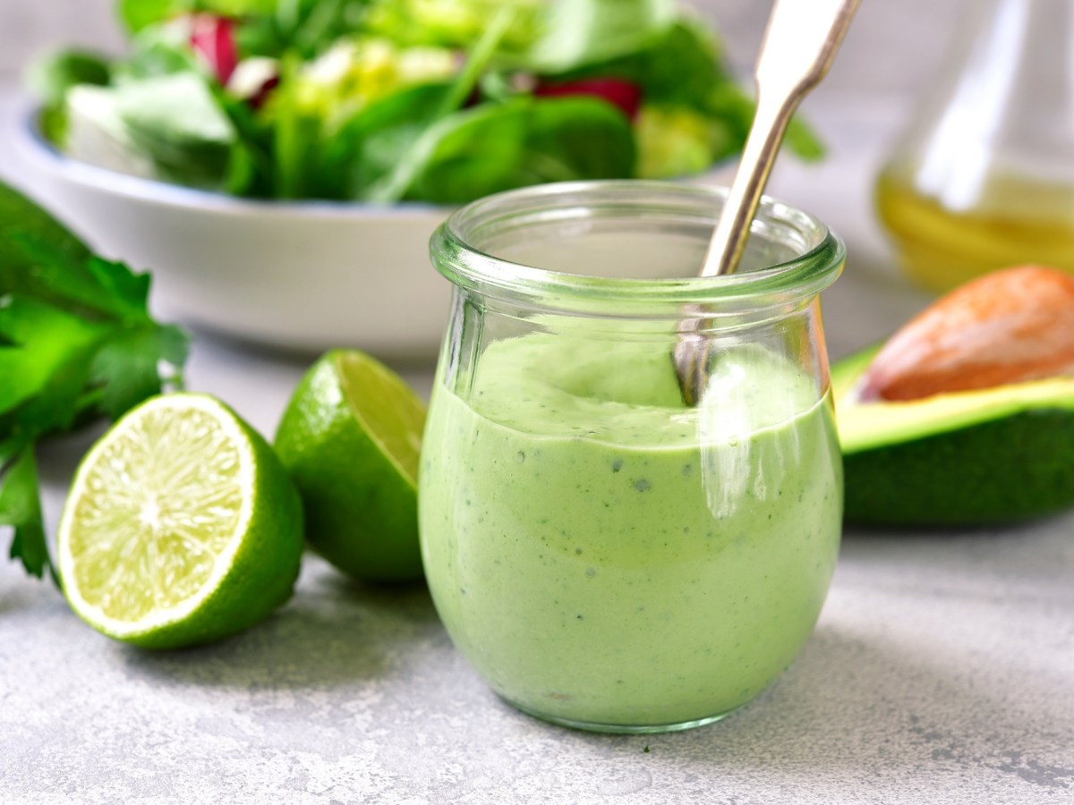 Ein Glas Avocado-Joghurt-Dressing mit Löffel, drumherum je eine halbierte Limette und Avocado.