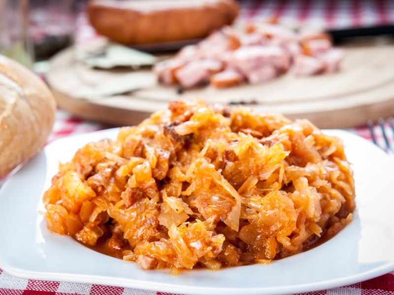 Eine Portion polnischer Eintopf Bigos auf einem Teller. Im Hintergrund ist eine Kabanossi zu sehen. Neben dem Teller liegt ein Brot.