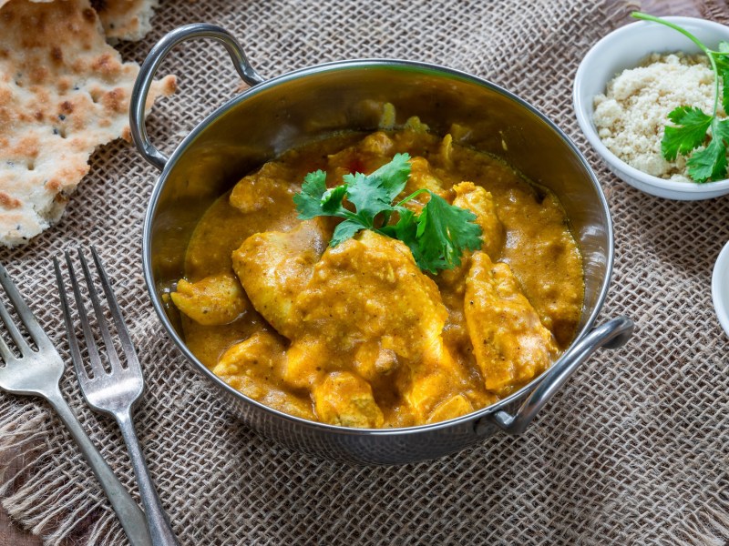 Chicken Korma in einer Metallschale mit Koriander garniert. Daneben liegen Naan-Brot, zwei Gabeln und eine Schale gemahlene Mandeln.