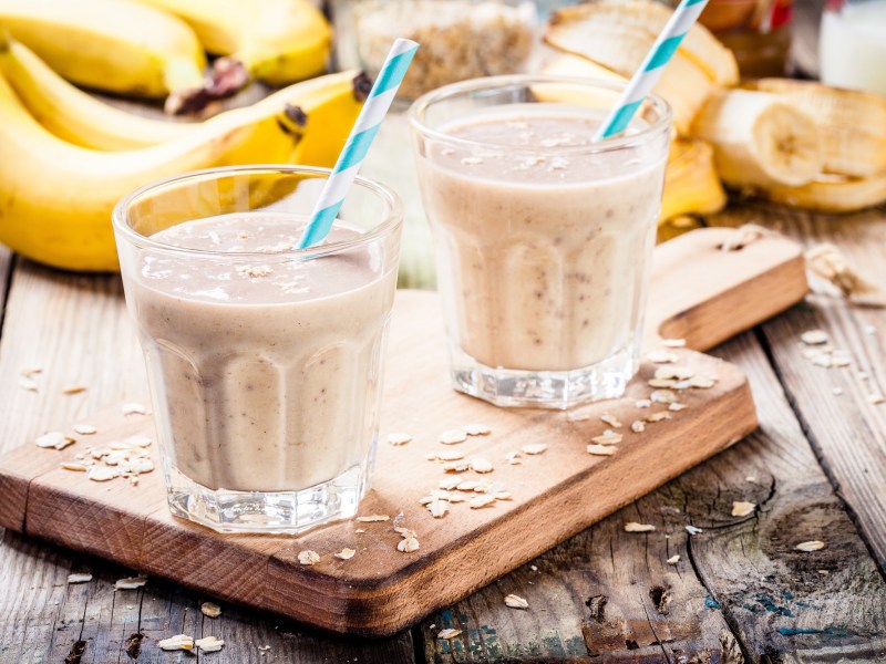 2 Gläser Erdnussbutter-Bananen-Smoothie mit Strohhalm und Haferflocken auf einem Brett, drumherum Bananen und Haferflocken.