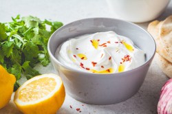 Eine Schüssel Feta-Knoblauch-Creme mit Öl und Chiliflocken garniert, daneben frische Kräuter und eine halbierte Zitrone sowie Fladenbrot.