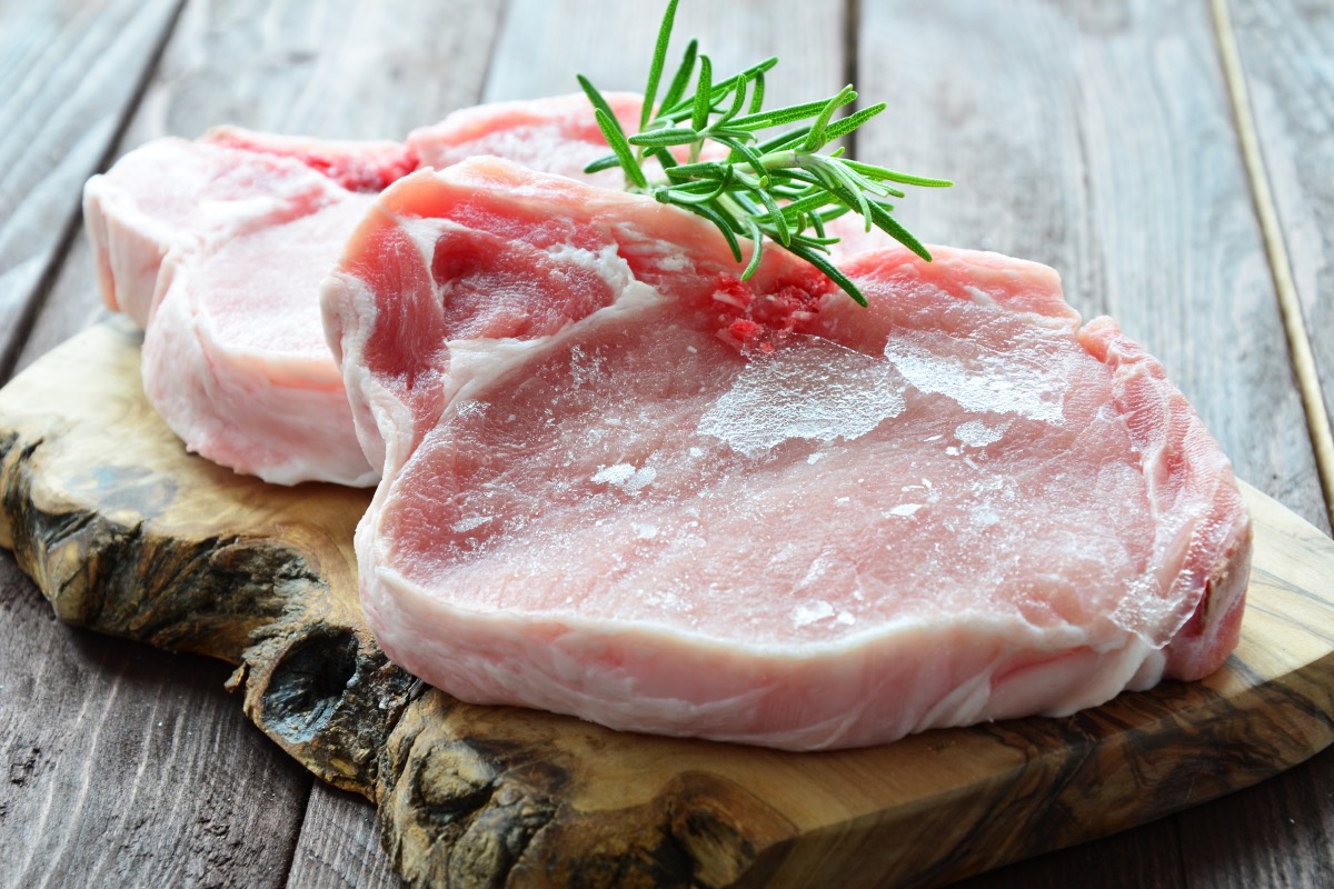 2 Schweine-Steaks auf einem Holzbrett, garniert mit einem Rosmarinzweig. Das Fleisch wurde eingefroren und aufgetaut.