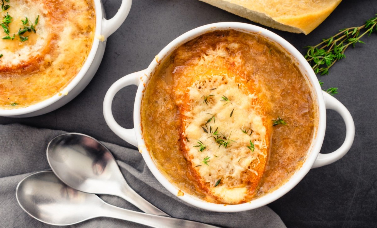 Eine Terrine mit französischer Zwiebelsuppe. Es befinden sich eine weitere Schüssel und zwei Suppenlöffel daneben.