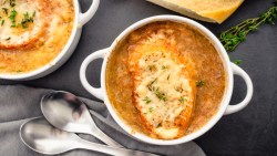 Eine Terrine mit französischer Zwiebelsuppe. Es befinden sich eine weitere Schüssel und zwei Suppenlöffel daneben.