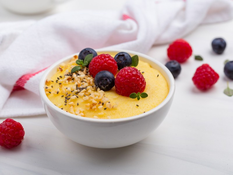 Eine weiße Schüssel Frühstücks-Polenta mit Nüssen und Beeren, drumherum ein Geschirrtuch und frische Beeren.