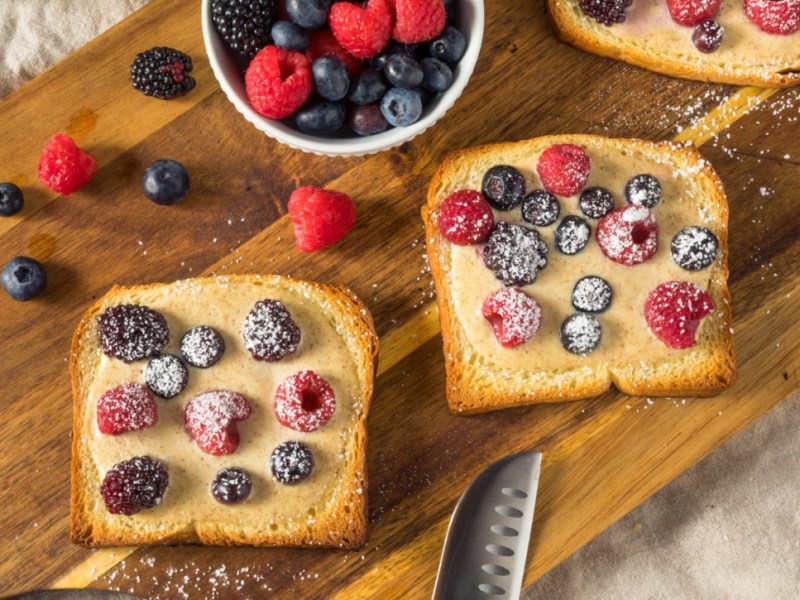 2 Scheiben Joghurt-Toast mit frischen Beeren auf einem Holzbrett. Daneben steht eine Schale mit Früchten und es liegt ein Messer am unteren Rand.