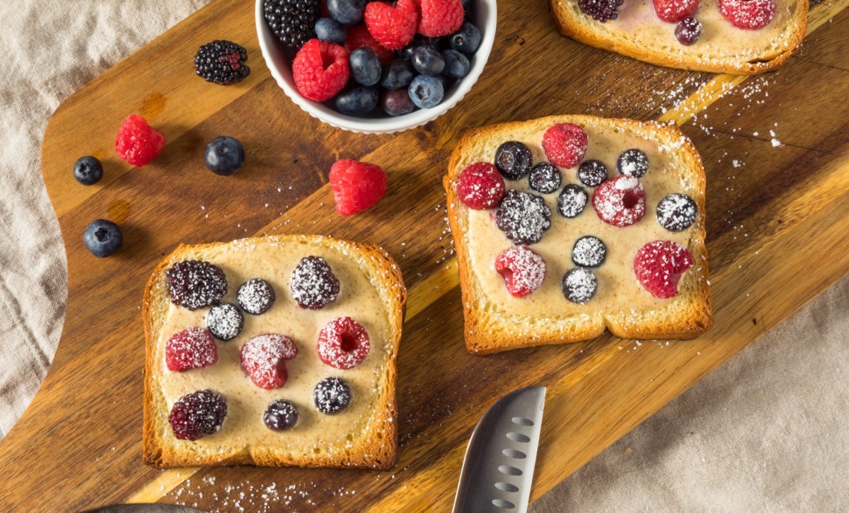 2 Scheiben Joghurt-Toast mit frischen Beeren auf einem Holzbrett. Daneben steht eine Schale mit Früchten und es liegt ein Messer am unteren Rand.