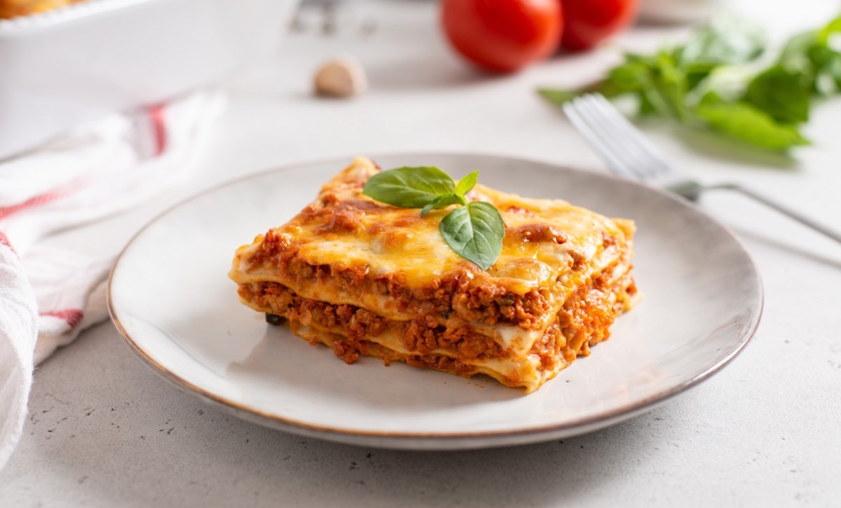 Ein Stück klassische Lasagne auf einem weißen Teller. Im Hintergrund sind frische Tomaten, Kräuter und die restliche Lasagne zu sehen.