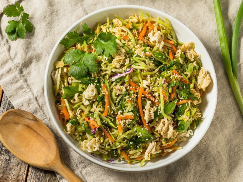 Knuspriger Ramen-Salat in einer weißen Schüssel, dekoriert mit etwas frischem Koriander. Daneben liegen ein Holzlöffel und eine Stange Frühlingszwiebel.