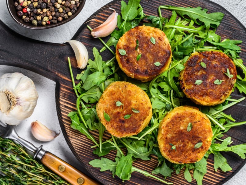 Vier Linsenfrikadellen auf einem Bett aus Rucola auf einem runden Holzbrett. Daneben steht eine Schale mit Pfeffer und es liegen Knoblauchzehen herum.