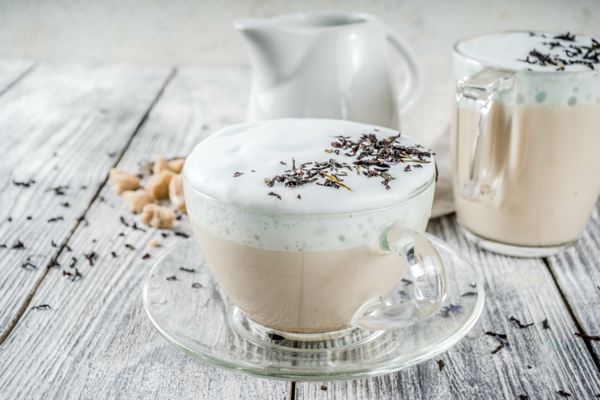 Durchsichtige Tasse London Fog Latte, Milchschaum und Lavendelblüten auf einem grauen Untergrund.