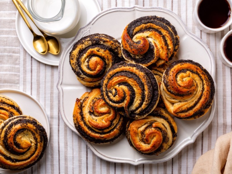 Ein Teller voller Mohnschnecken, von oben fotografiert. Daneben stehen zwei Tassen Kaffee und ein weiterer Teller mit Mohnschnecken. Außerdem ein Kännchen Milch.