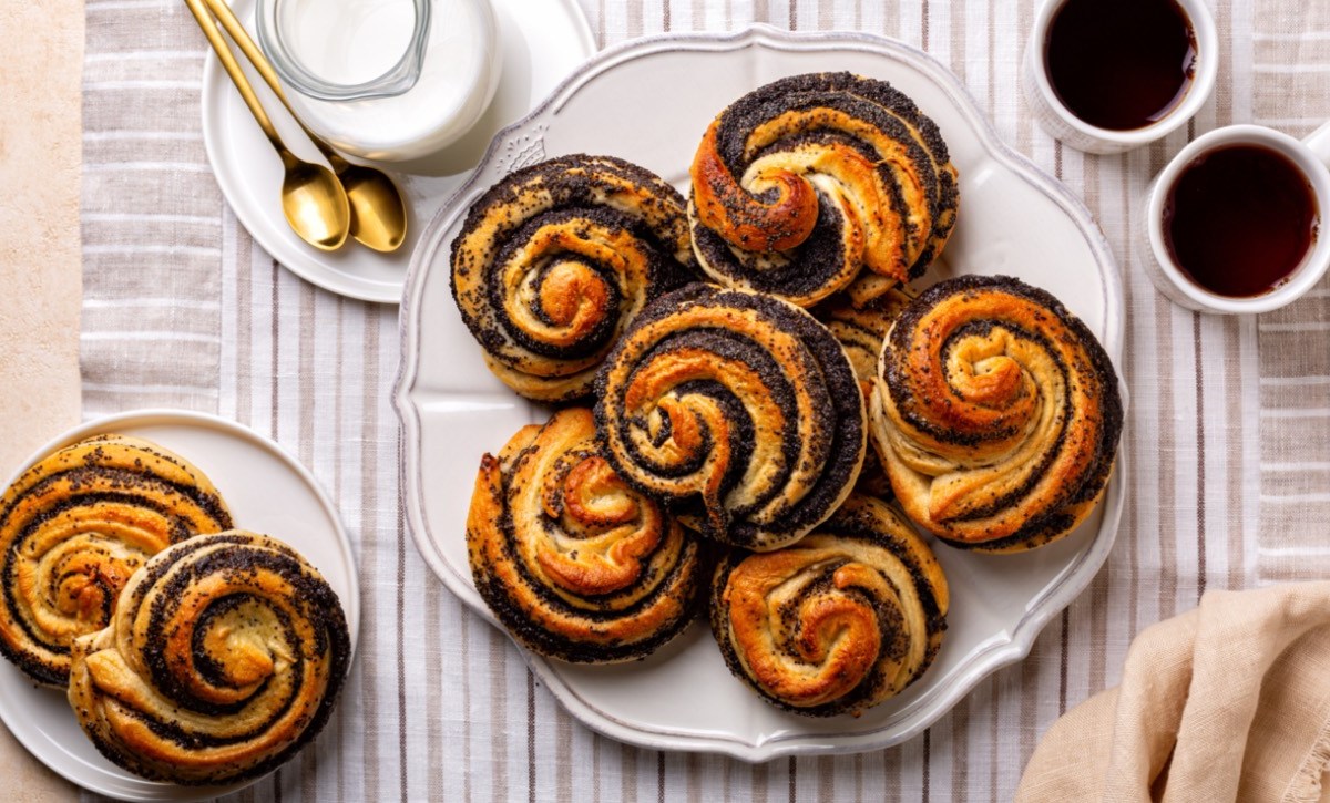 Ein Teller voller Mohnschnecken, von oben fotografiert. Daneben stehen zwei Tassen Kaffee und ein weiterer Teller mit Mohnschnecken. Außerdem ein Kännchen Milch.