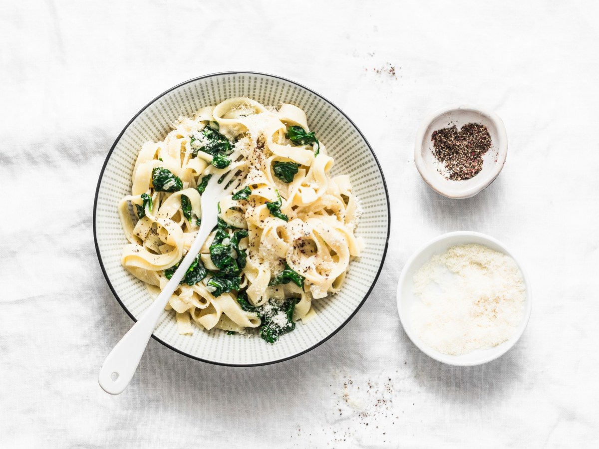 Ein Teller Nudeln mit Spinat-Käse-Soße mit Gabel in der Draufsicht, daneben je eine Schüssel mit Pfeffer und geriebenem Parmesan.