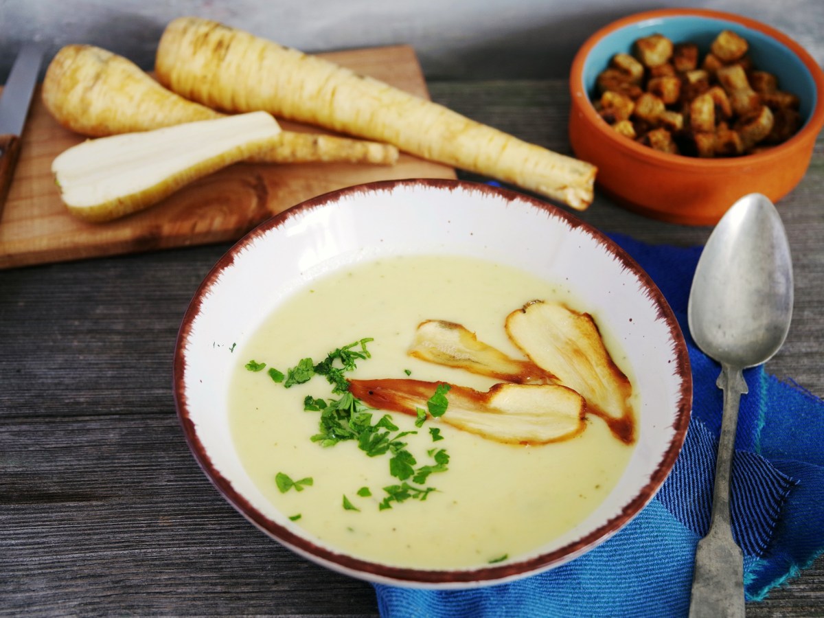 Pastinakensuppe in einer weißen Schüssel mit Pastinakenchips und Petersilie garniert. Daneben liegen Croutons und ganze Pastinaken.