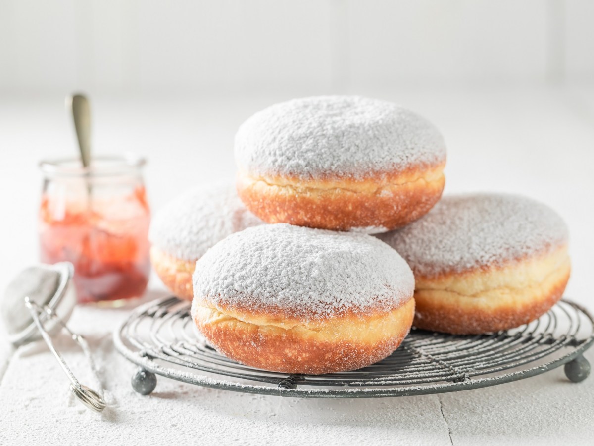 Mehrere polnische Krapfen auf einem Kucnengitter gestapelt, bestreut mit Puderzucker, daneben ein Sieb und ein Glas Marmelade.