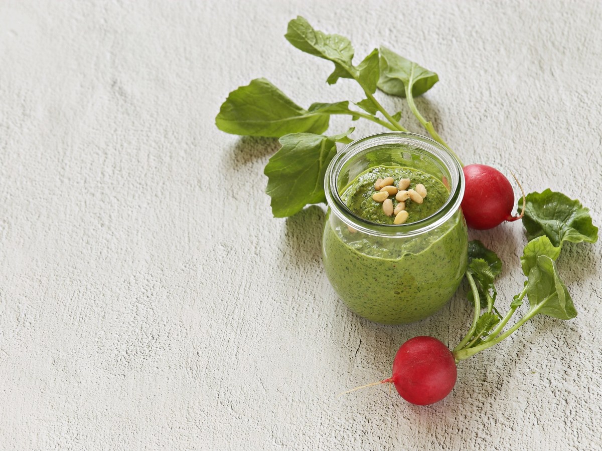 Ein Glas mit grünem Pesto und Radieschen daneben auf einem weißen Untergrund