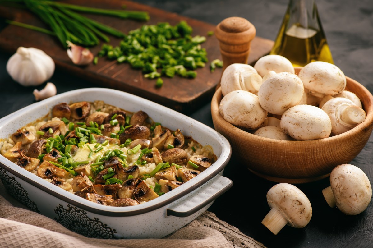 Reisauflauf mit Pilzen in Auflaufform, daneben eine Schale mit Champignons und Kräuter.