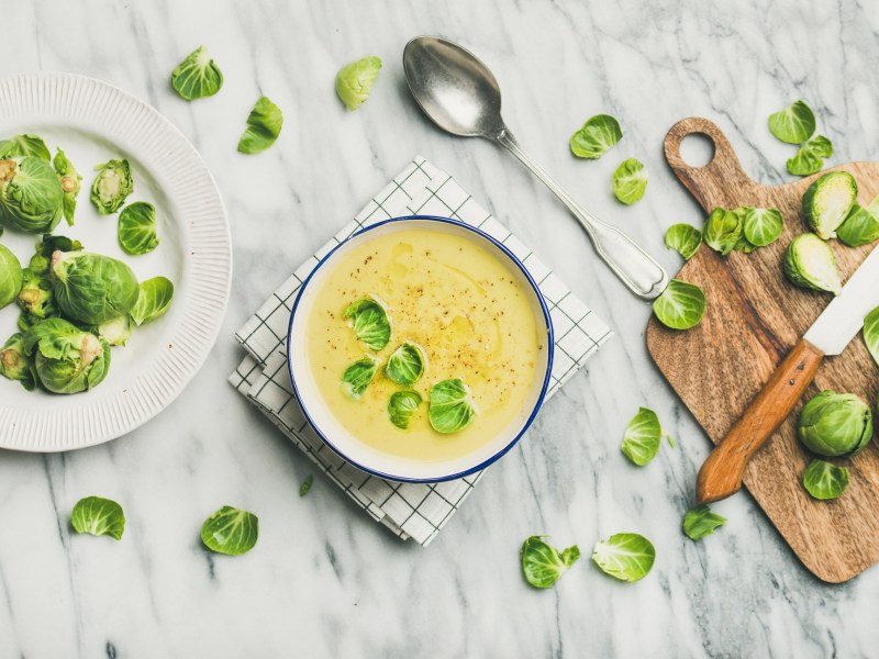 Eine Schüssel cremige Rosenkohlsuppe mit Schneidebrett, Messer, Rosenkohl und Löffel.