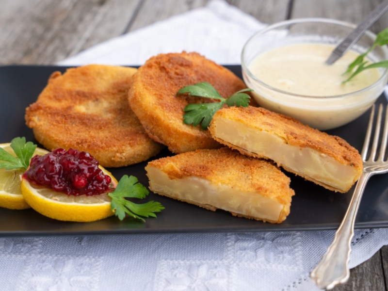 Mehrere Sellerieschnitzel auf einem schwarzen, rechteckigen Teller. Mit auf dem Teller sind zwei Zitronenscheiben, eine davon mit Preiselbeermarmelade, und ein Schälchen mit weißer Soße.