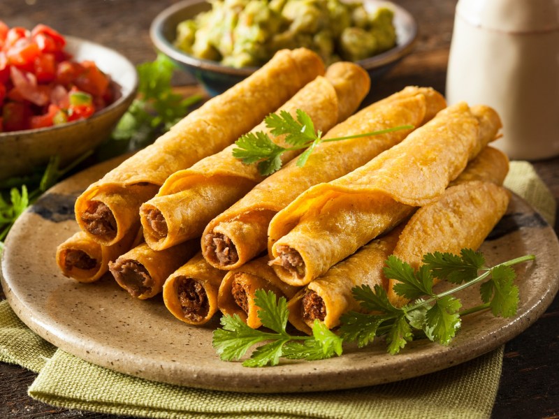 Taquitos mit Hackfleischfüllung auf einem beigen Teller, garniert mit Koriander und serviert mit Tomaten-Salsa, Guacamole und Sour Cream.