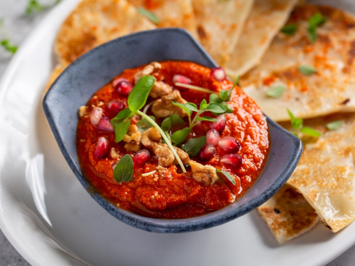 Eine Schale Tomate-Walnuss-Aufstrich, garniert mit Walnüssen, Kräutern und Granatapfelkernen, auf einem Teller, auf dem auch dreieckige Fladenbrote liegen, Nahaufnahme.