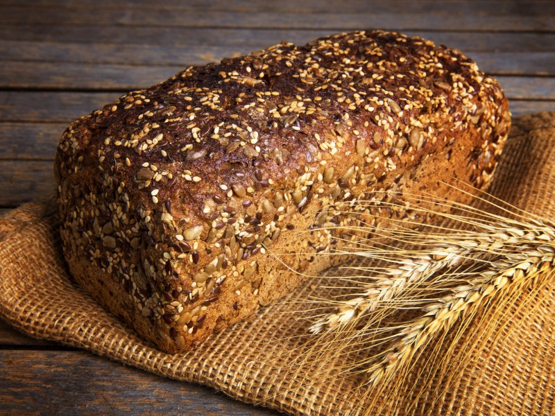 Ein Laib Vollkornbrot mit verschiedenen Saaten auf einem Mehlsack. Daneben zwei Getreideähren.