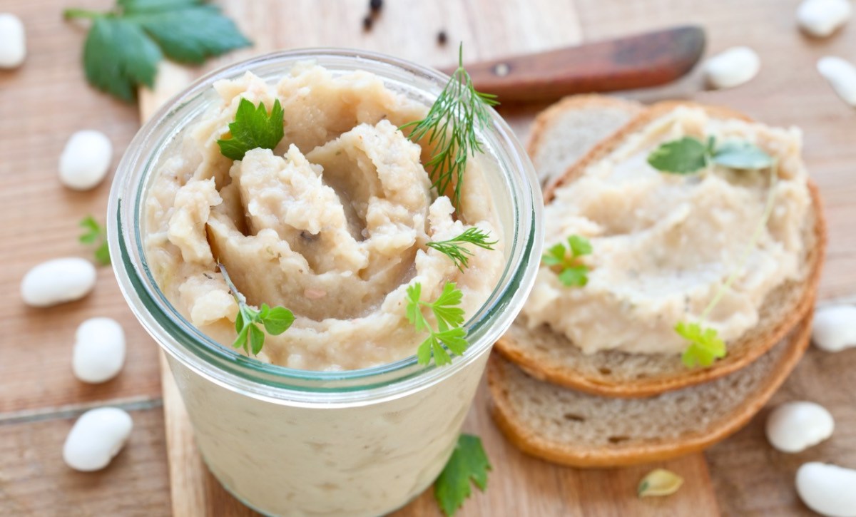 Weiße-Bohnen-Aufstrich im Glas. Daneben eine Scheibe Brot mit Aufstrich.