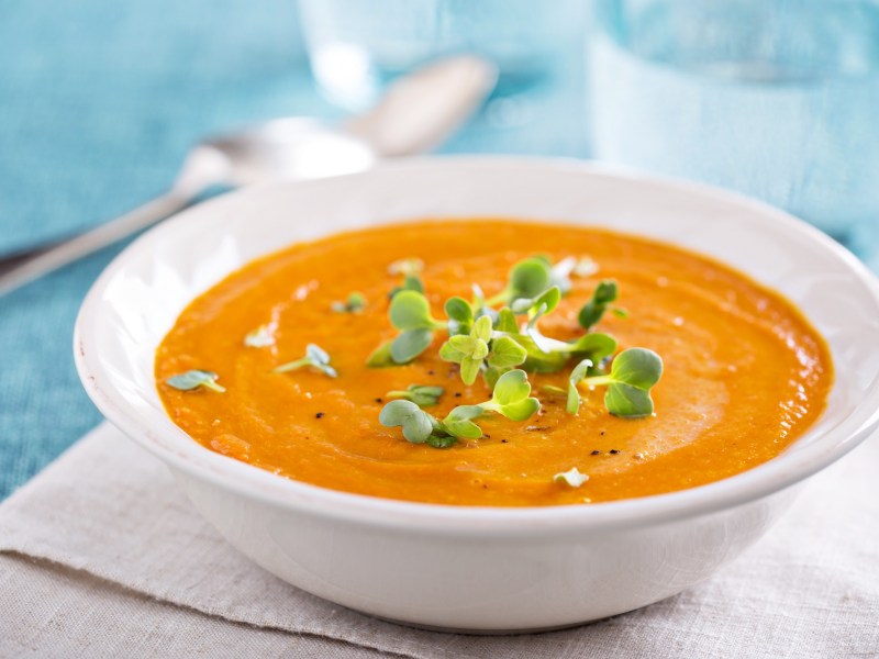 Ein tiefer Teller Zucchini-Möhren-Suppe garniert mit Kräutern auf einem Platzdeckchen, Dahinter unscharf ein Glas Wasser, Nahaufnahme.